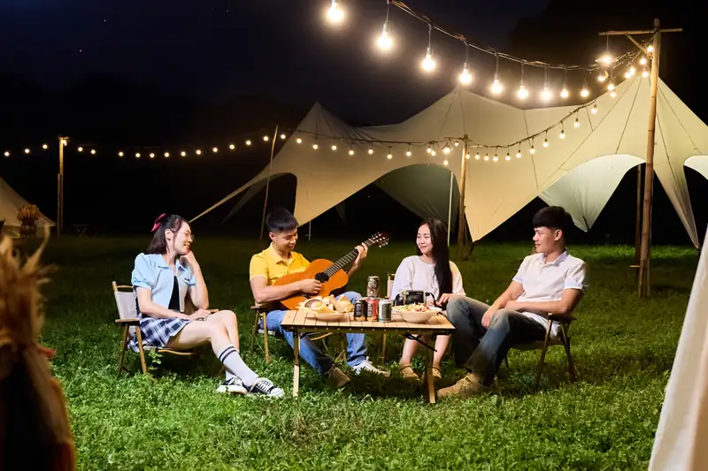 Nhóm khách đàn hát bên bàn dã ngoại dưới mái bạt buổi tối – Serena Camping