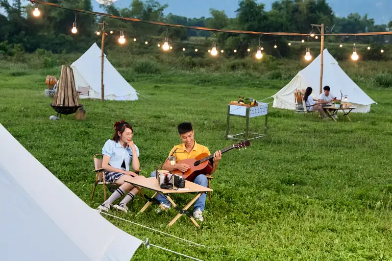 Khách cắm trại trong ngày ngồi đàn hát bên lều trên bãi cỏ có dây đèn ở Serena Kim Bôi