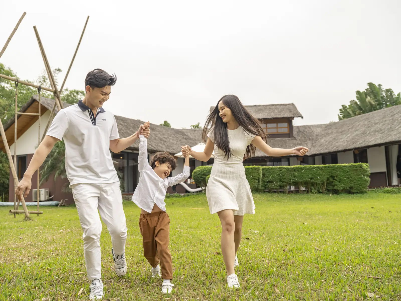 Bố mẹ nắm tay con nhỏ chạy trên bãi cỏ trước dãy Bungalow mái tranh ở Serena Kim Bôi